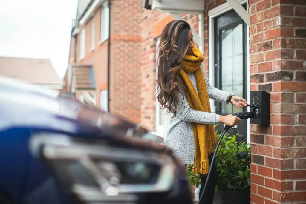 Woman in a yellow scarf plugging her electric car into a wall charger beside her front door