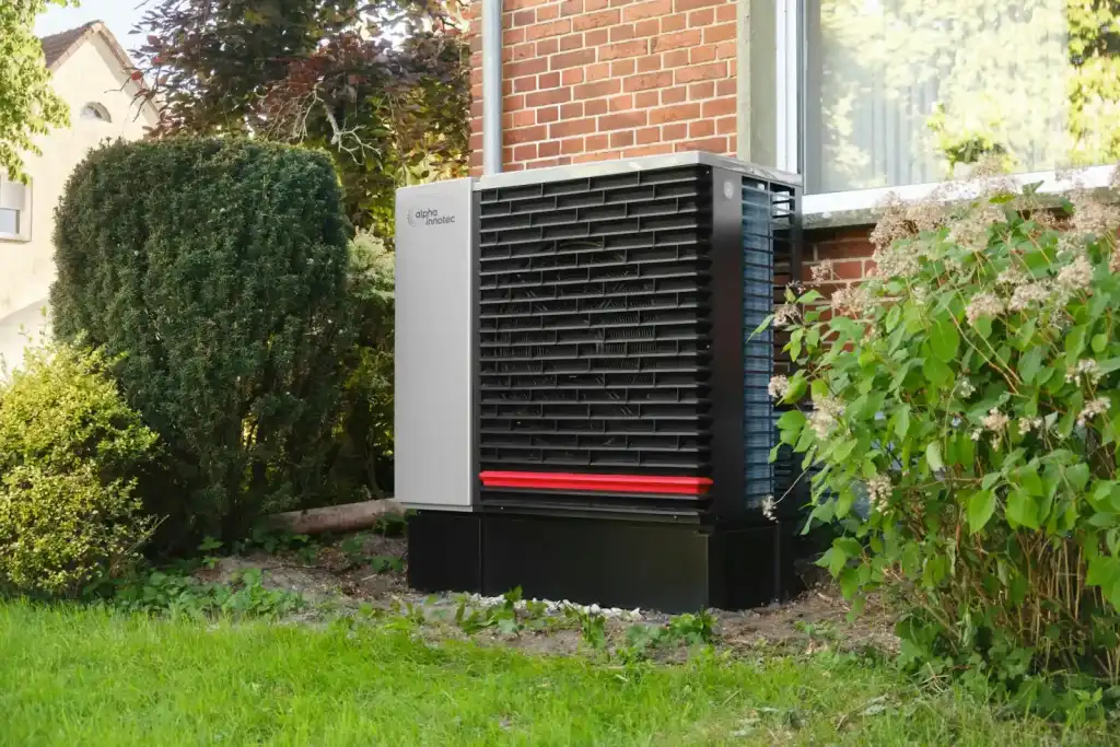 Outdoor heat pump unit on the lawn beside a brick house, framed by green shrubs
