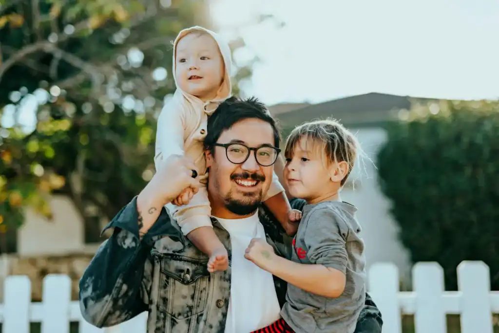 Smiling father holding a toddler on his shoulders and hugging his young son outside a home with a white picket fence