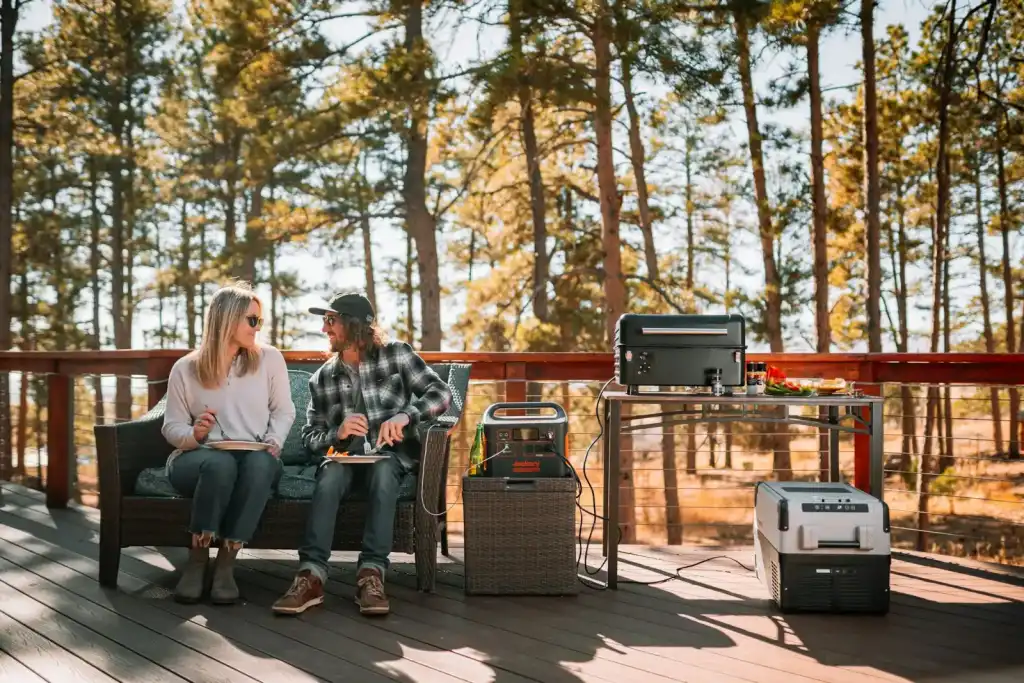 Couple relaxing on a sunny deck among pine trees with a portable power station running a grill and a cooler